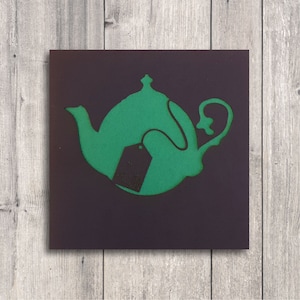 May include: A green teapot with a black tea bag hanging from the spout. The teapot is on a dark brown background.