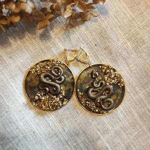 May include: A pair of round earrings with a gold-colored border. Each earring features a detailed bronze-colored snake design on a dark background, accented with gold-colored embellishments. The earrings are displayed on a neutral linen surface.