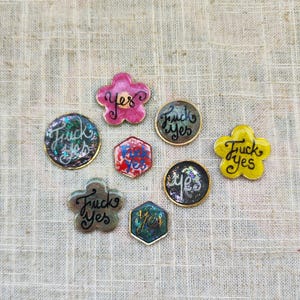 Può includere: Una collezione di spille fatte a mano in varie forme, tra cui fiori, cerchi e un esagono. Ogni spilla presenta le parole "Fuck Yes" o "Yes" in un carattere corsivo. Le spille hanno un bordo dorato metallico e sfondi colorati e iridescenti.