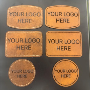 May include: Six brown faux leather patches in various shapes including rectangular, rounded rectangular, and circular. Each patch features the text YOUR LOGO HERE engraved in black. The patches have visible stitching along the edges in either white or black thread.