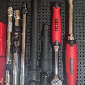 Op de afbeelding: Een verzameling gereedschappen voor monteurs, waaronder ratels, verlengstukken en doppen, netjes gerangschikt in een zwarte opbergbak. De gereedschappen hebben zilverkleurige metalen lichamen en rode handvatten met het "ICON" logo.
