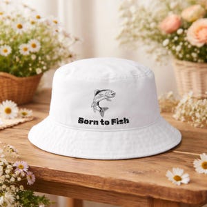 Könnte beinhalten: Weißer Fischerhut mit einer schwarzen Grafik eines springenden Fisches und dem Text "Born to Fish". Der Hut ist aus einem weichen Material gefertigt und eignet sich für Outdoor-Aktivitäten. Der Hut liegt auf einer Holzoberfläche.