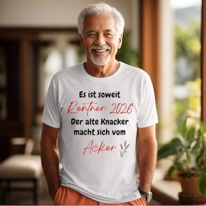 Könnte beinhalten: Ein weißes T-Shirt mit schwarzem und rotem Text, der lautet: "Es ist soweit Rentner 2026 Der alte Knacker macht sich vom Acker". Das T-Shirt wird von einer lächelnden Person getragen.