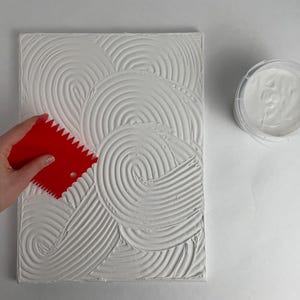 Può includere: Una tela bianca strutturata con un motivo a spirale viene lavorata con una cazzuola rossa dentata. Un contenitore di pasta bianca è accanto alla tela. L'immagine mostra un progetto artistico fai-da-te.