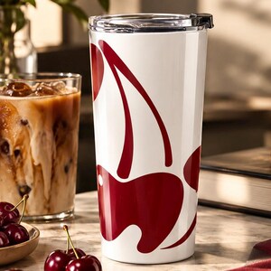 May include: A white cylindrical travel tumbler featuring a large, stylized dark red cherry graphic with a long stem. The tumbler has a clear plastic lid with a black sliding closure mechanism. The design is smooth and glossy, wrapping around the exterior.