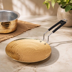 May include: A round, flat, hammered gold-coloured metal pan with a long, dual-wire handle featuring black plastic grips. The surface of the pan has a textured, dimpled finish, and the handle is securely attached with two visible metal rivets.