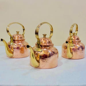 Op de afbeelding: Drie gehamerde koperen theeketels met handvatten en tuiten van gepolijst messing. Elke ketel heeft een ronde behuizing met een getextureerd, gedeukt oppervlak, een bijpassend deksel met een knop en een gebogen handvat aan de bovenkant.