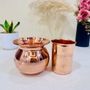 May include: Two copper vessels sit on a white surface. One is a small, round pot with a wide rim. The other is a cylindrical cup with etched floral designs. The vessels have a polished, reflective surface.