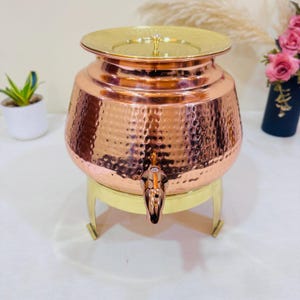 May include: A copper beverage dispenser with a hammered finish, brass base, and lid. The dispenser features a tap for dispensing and a decorative handle. The copper has a reddish-brown colour, and the design is ornate. The dispenser is suitable for serving drinks.