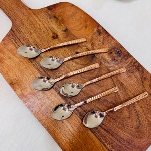 May include: A set of six small spoons with polished silver-tone bowls and textured copper-tone handles. Each spoon features a distinct hammered pattern along the length of the handle, providing a tactile finish to the metallic design.
