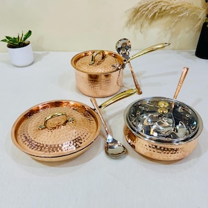 May include: A set of hammered copper cookware including a lidded saucepan with a long handle, a lidded frying pan, and a lidded serving bowl with a glass lid. Two matching metal ladles with long handles are included with the copper kitchenware set.