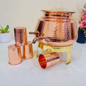 Könnte beinhalten: Ein Kupfer-Wasserspender mit Zapfhahn, auf einem Messingständer, und vier passende Kupferbecher. Der Spender hat eine gehämmerten Textur. Die Becher haben ein Blumenmuster.