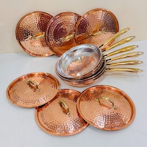 Pode incluir: Um conjunto de utensílios de cozinha em cobre martelado com várias frigideiras redondas com pegas douradas brilhantes e tampas circulares a condizer. O interior das frigideiras tem um acabamento metálico prateado, enquanto o exterior e as tampas apresentam uma superfície de cobre martelado texturizado com pegas douradas polidas.