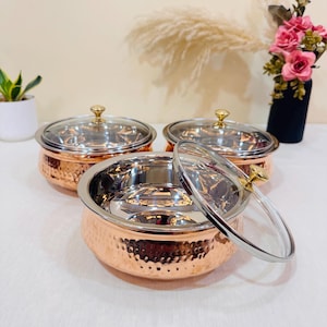 May include: Three copper-colored serving bowls with glass lids and gold-colored knobs. The bowls have a hammered texture on the exterior and a shiny, silver interior. One lid is partially removed, revealing the interior. The bowls are arranged on a white surface.