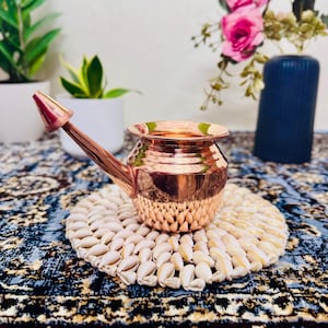 May include: A copper watering can with a long spout and a rounded body. It rests on a round mat made of small, white shells. The background features plants and a blue vase with pink flowers. The can is likely used for watering plants.