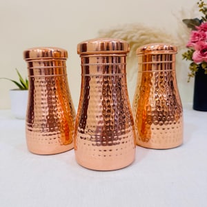 May include: Three copper-colored, hammered-finish water bottles with matching lids. The bottles have a bulbous shape and a textured surface. The lids are flat and round. The bottles are arranged in a group, showcasing their design and material.