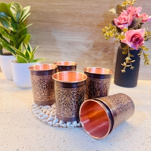 Puede incluir: Juego de cinco vasos para beber de color cobre. Los vasos tienen una banda marrón oscuro con un patrón ornamental y ondulado. El interior de los vasos es de color cobre brillante. Un vaso está tumbado de lado. Los vasos están sobre una superficie blanca texturizada.