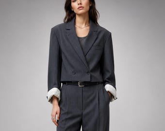 May include: A dark gray cropped blazer with long sleeves and white cuffs, paired with matching high-waisted wide-leg trousers. The blazer features a double-breasted front closure, and the trousers are cinched at the waist with a thin black belt.