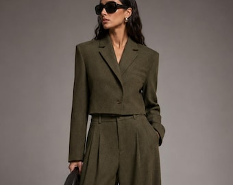 May include: An olive green two-piece set consisting of a cropped blazer with a notched lapel and a single button closure, paired with high-waisted, wide-leg trousers featuring front pleats. The outfit is styled with black sunglasses and a black bag.