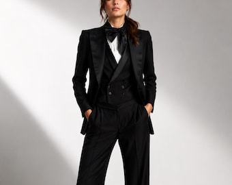 May include: A formal black suit with a satin lapel tuxedo jacket, vest, and wide-leg trousers. The outfit includes a white dress shirt and a black bow tie. The model is standing with hands in pockets.