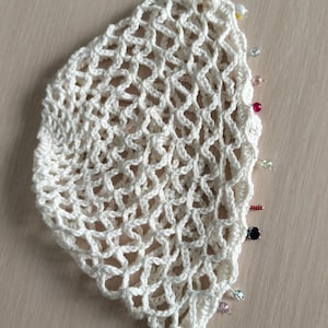 May include: A semi-circular off-white crochet doily featuring an openwork net pattern. The curved edge is finished with a series of small, multi-colored glass beads in shades of red, blue, pink, and clear, attached individually along the border.