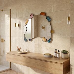 May include: An irregular-shaped wall mirror featuring a thin metal frame accented with various colorful, polished stone-like pieces in shades of blue, orange, yellow, and white, mounted on a light-colored tiled wall above a wooden vanity.
