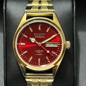 May include: A gold-toned Citizen Automatic watch with a vibrant red dial. The watch features a gold band and a date display showing "SAT 5." The watch face includes the text "CITIZEN AUTOMATIC" and "21 JEWELS."