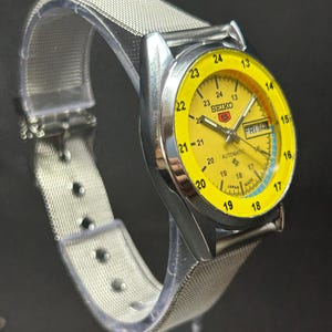 Puede incluir: Un reloj autom&aacute;tico Seiko 5 vintage con esfera amarilla y caja de acero inoxidable plateado. El reloj tiene una pantalla de d&iacute;a y fecha y una correa de malla plateada. La esfera tiene n&uacute;meros y marcadores negros.