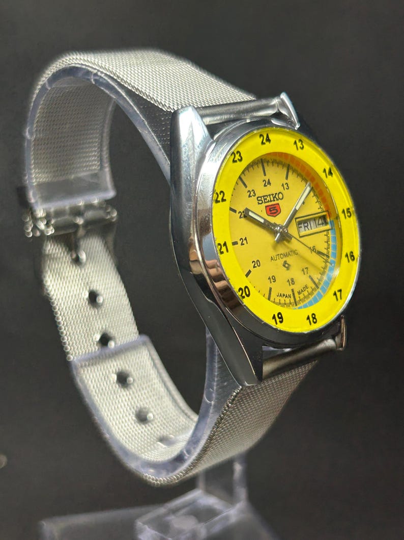 Puede incluir: Un reloj autom&aacute;tico Seiko 5 vintage con esfera amarilla y caja de acero inoxidable plateado. El reloj tiene una pantalla de d&iacute;a y fecha y una correa de malla plateada. La esfera tiene n&uacute;meros y marcadores negros.