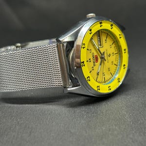 Puede incluir: Un reloj Seiko plateado con esfera amarilla y correa de malla. La esfera muestra el d&iacute;a y la fecha, con n&uacute;meros y agujas negros. El reloj est&aacute; en &aacute;ngulo, mostrando sus detalles sobre un fondo oscuro.