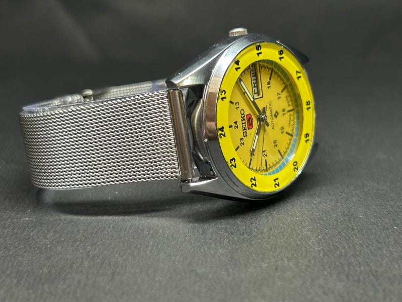 Puede incluir: Un reloj Seiko plateado con esfera amarilla y correa de malla. La esfera muestra el d&iacute;a y la fecha, con n&uacute;meros y agujas negros. El reloj est&aacute; en &aacute;ngulo, mostrando sus detalles sobre un fondo oscuro.