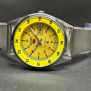 Puede incluir: Un reloj de pulsera autom&aacute;tico Seiko plateado con una esfera amarilla brillante. El reloj tiene una correa de malla y una pantalla de d&iacute;a y fecha. La esfera tiene n&uacute;meros y marcadores negros, con el d&iacute;a mostrado como "FRI 14".
