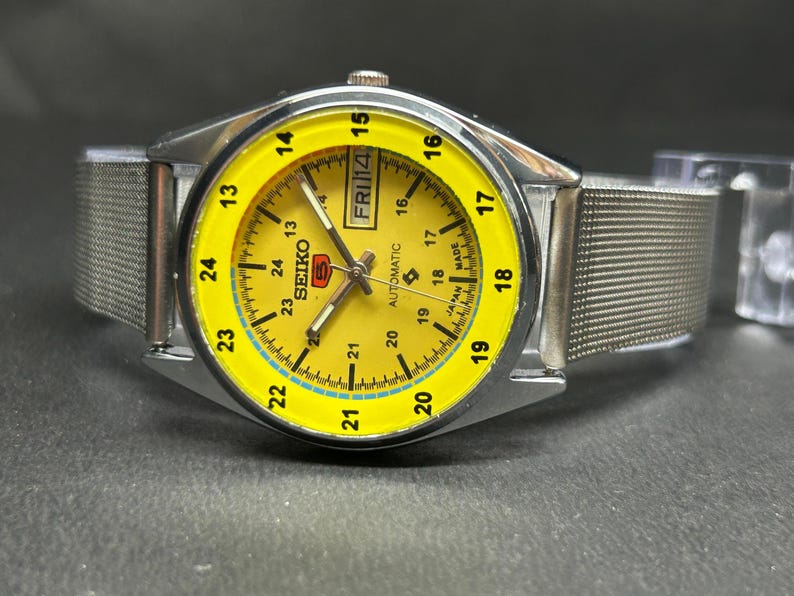 Puede incluir: Un reloj de pulsera autom&aacute;tico Seiko plateado con una esfera amarilla brillante. El reloj tiene una correa de malla y una pantalla de d&iacute;a y fecha. La esfera tiene n&uacute;meros y marcadores negros, con el d&iacute;a mostrado como "FRI 14".