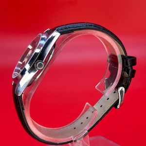 May include: A silver-toned wristwatch with a black leather band. The watch face is visible, and the band is secured with a silver buckle. The watch is displayed on a clear stand against a red background.