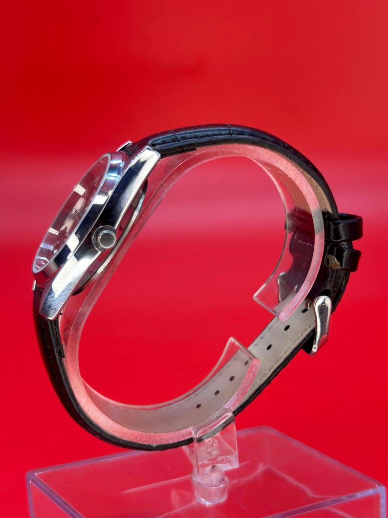 May include: A silver-toned wristwatch with a black leather band. The watch face is visible, and the band is secured with a silver buckle. The watch is displayed on a clear stand against a red background.