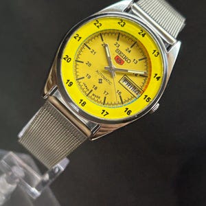 Puede incluir: Un reloj de pulsera autom&aacute;tico Seiko vintage con esfera amarilla brillante y correa de malla plateada. La esfera muestra el d&iacute;a y la fecha, con un indicador de 24 horas. Fabricado en Jap&oacute;n.