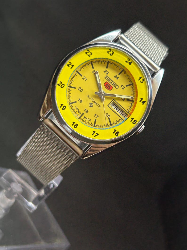 Puede incluir: Un reloj de pulsera autom&aacute;tico Seiko vintage con esfera amarilla brillante y correa de malla plateada. La esfera muestra el d&iacute;a y la fecha, con un indicador de 24 horas. Fabricado en Jap&oacute;n.