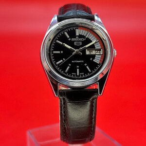 May include: A silver-toned Seiko automatic wristwatch with a black dial and a black leather band. The watch face features white markers, a day-date display, and the Seiko logo. The watch is displayed against a red background.