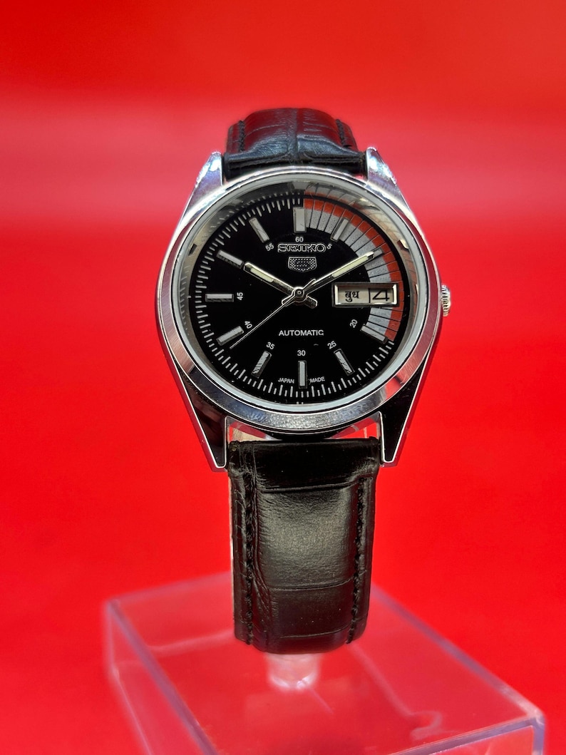 May include: A silver-toned Seiko automatic wristwatch with a black dial and a black leather band. The watch face features white markers, a day-date display, and the Seiko logo. The watch is displayed against a red background.