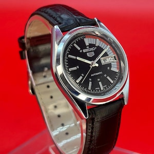 May include: A vintage Seiko automatic watch with a black dial and silver-tone case. The watch features a black leather band and a date display. The dial includes the Seiko logo and the word "AUTOMATIC". The watch is set against a red background.