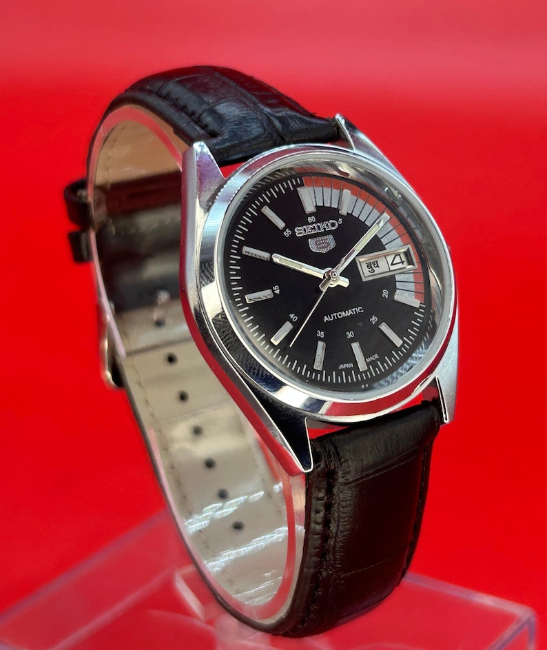 May include: A vintage Seiko automatic watch with a black dial and silver-tone case. The watch features a black leather band and a date display. The dial includes the Seiko logo and the word "AUTOMATIC". The watch is set against a red background.