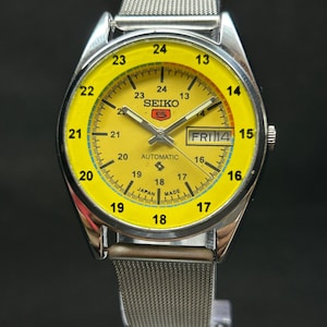 Puede incluir: Un reloj de pulsera autom&aacute;tico Seiko 5 vintage con una esfera de color amarillo brillante. El reloj tiene una caja plateada y una correa de malla met&aacute;lica. La esfera muestra el d&iacute;a y la fecha, con el d&iacute;a que indica "FRI 14". El reloj tiene una escala de 24 horas.