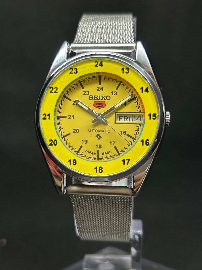 Puede incluir: Un reloj de pulsera autom&aacute;tico Seiko 5 vintage con una esfera de color amarillo brillante. El reloj tiene una caja plateada y una correa de malla met&aacute;lica. La esfera muestra el d&iacute;a y la fecha, con el d&iacute;a que indica "FRI 14". El reloj tiene una escala de 24 horas.