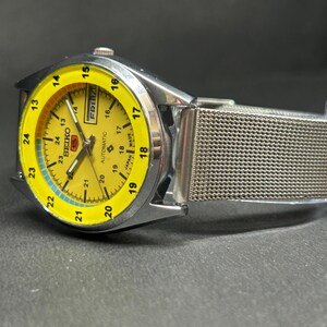 Puede incluir: Un reloj de pulsera autom&aacute;tico Seiko con esfera amarilla y correa de malla plateada. La esfera muestra el d&iacute;a y la fecha, con un indicador de tiempo de 24 horas. El reloj tiene una caja de acero inoxidable.