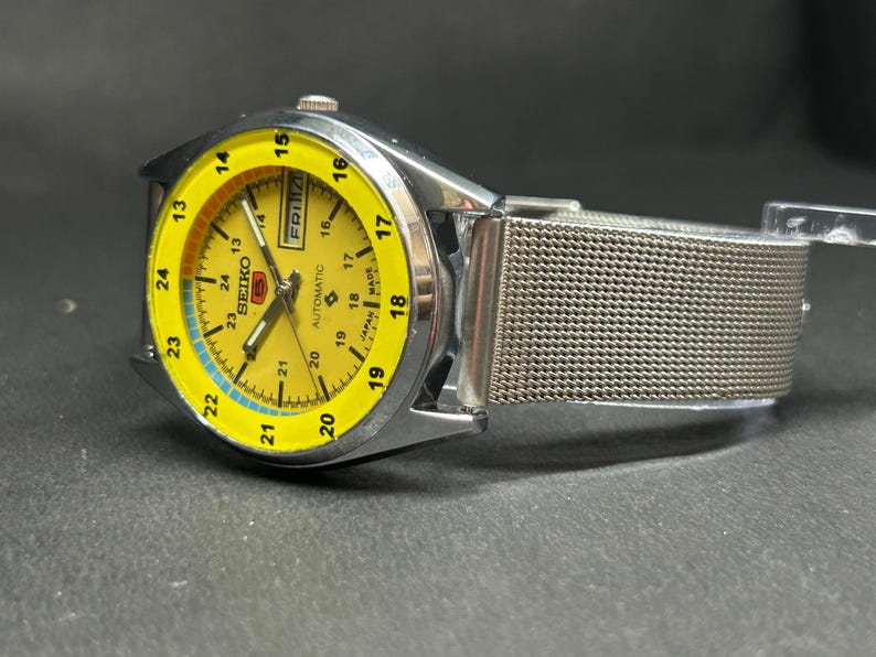Puede incluir: Un reloj de pulsera autom&aacute;tico Seiko con esfera amarilla y correa de malla plateada. La esfera muestra el d&iacute;a y la fecha, con un indicador de tiempo de 24 horas. El reloj tiene una caja de acero inoxidable.