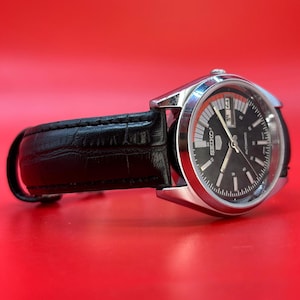 May include: A silver-toned Seiko automatic watch with a black dial and a black leather band. The watch face has white and orange accents, with a day and date display. The watch is set against a red background.