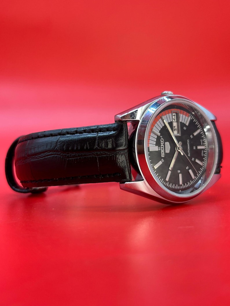 May include: A silver-toned Seiko automatic watch with a black dial and a black leather band. The watch face has white and orange accents, with a day and date display. The watch is set against a red background.