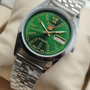 May include: A silver-toned Seiko automatic wristwatch with a green dial. The watch features gold-colored hands and hour markers, a day-date display, and a stainless steel band. The dial displays the text "SEIKO" and "AUTOMATIC".