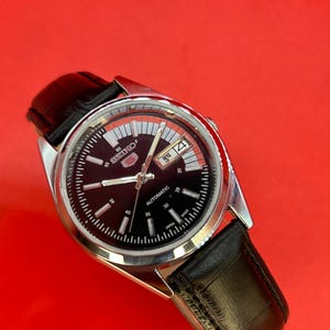 May include: A Seiko 5 automatic wristwatch with a black dial and a silver-tone case. The watch features a day and date display and a black leather strap. The watch face has white hour markers and hands.