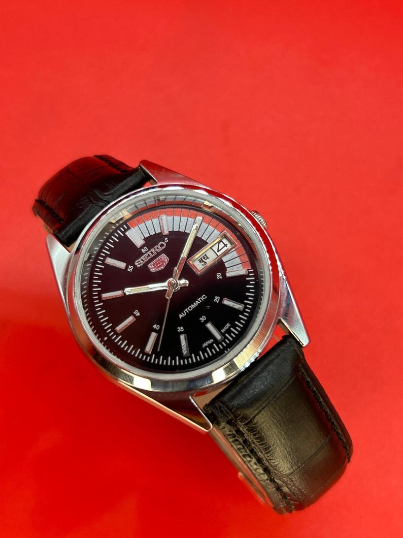 May include: A Seiko 5 automatic wristwatch with a black dial and a silver-tone case. The watch features a day and date display and a black leather strap. The watch face has white hour markers and hands.
